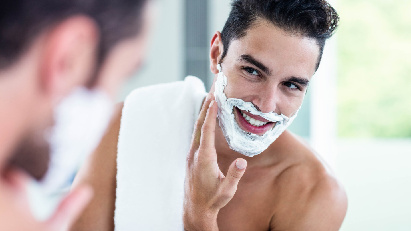 Richtig rasieren, Rasurbrand vermeiden: Junger Mann mit Rasierschaum im Gesicht schaut in den Spiegel.
